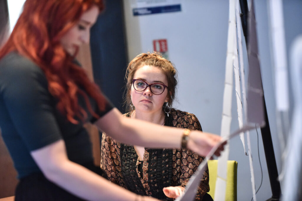 A performer examining a connected string of punchcards, whilst another performer looks at them