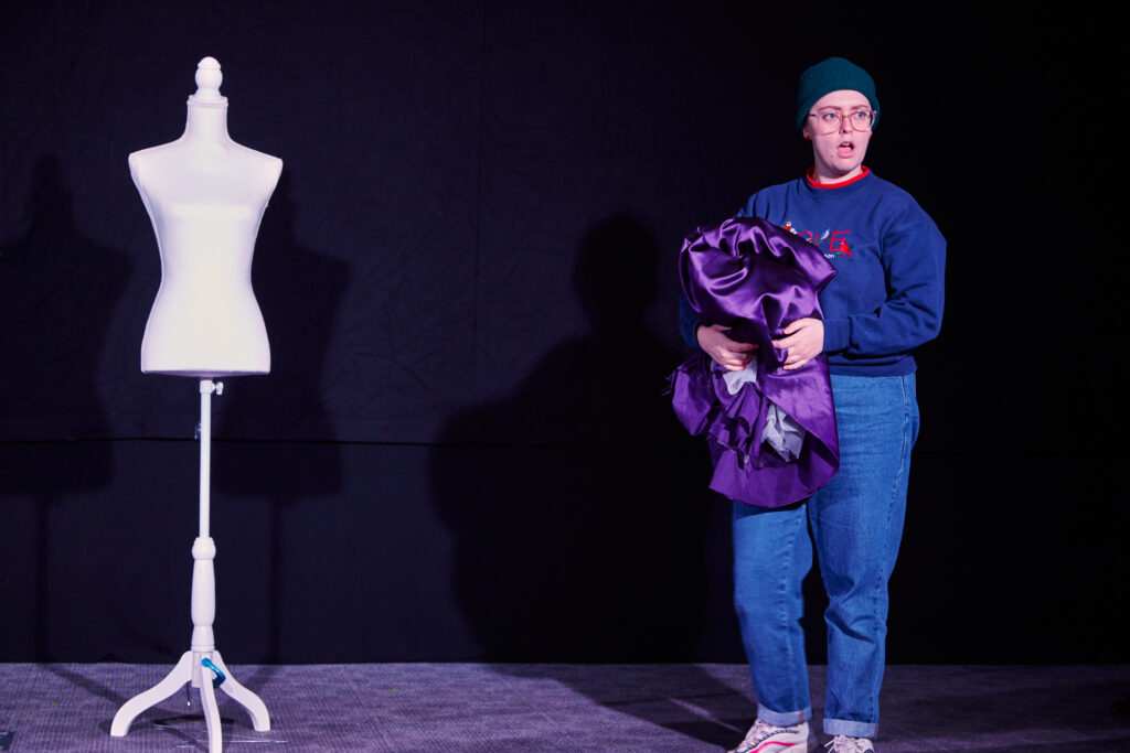 A performer wearing a beanie hat, holding shiny purple fabric, standing next to a naked dressmakers' doll